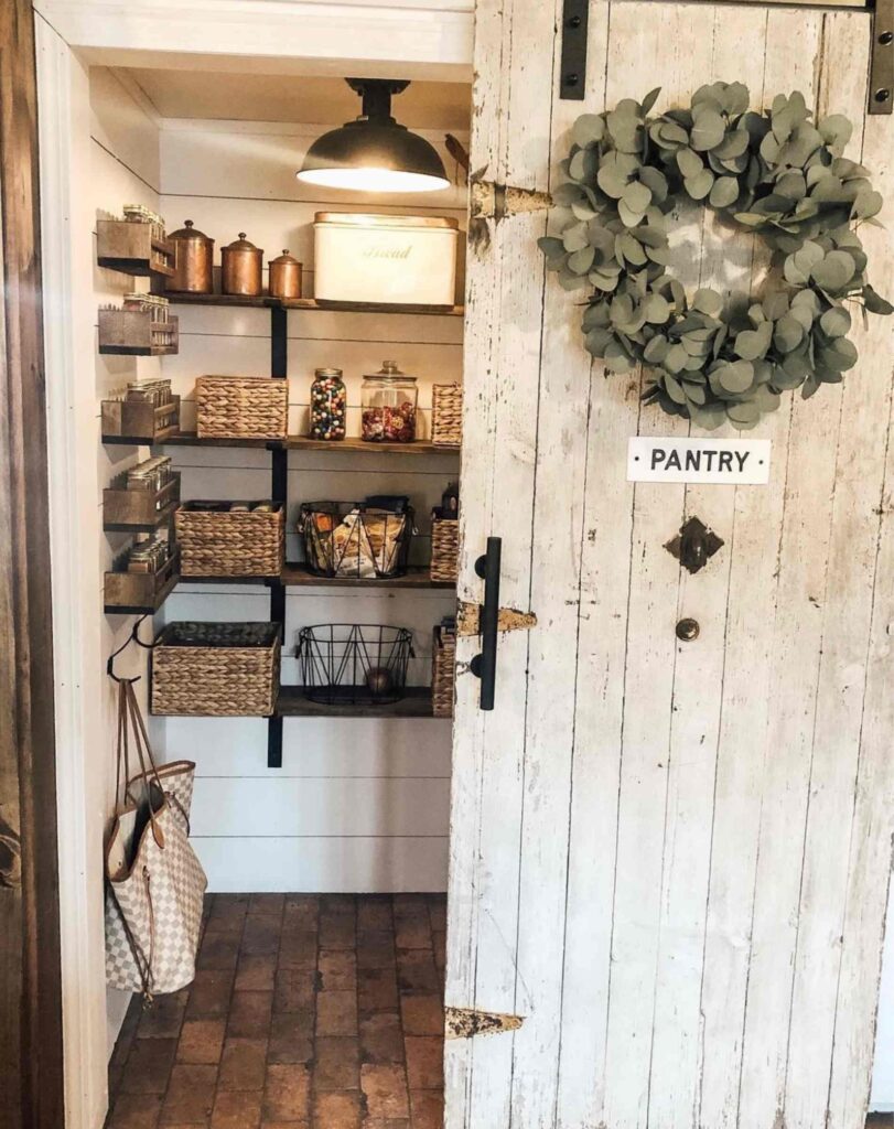 A rustic pantry with a sliding farmhouse door, bins, spices on display, wire baskets, glass jars, and a bread bin.