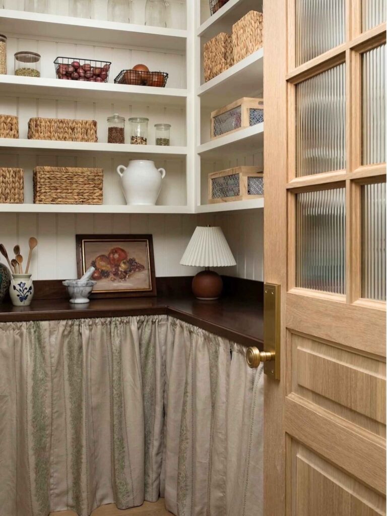 Country pantries with open shelving, lamp, woven bins, jars, and painting.