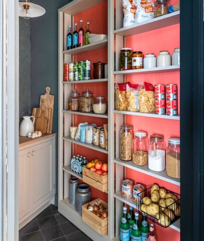 Country pantry with a pink pop of color, storing pantry staples, jars, wood cutting boards, and bins with produce.