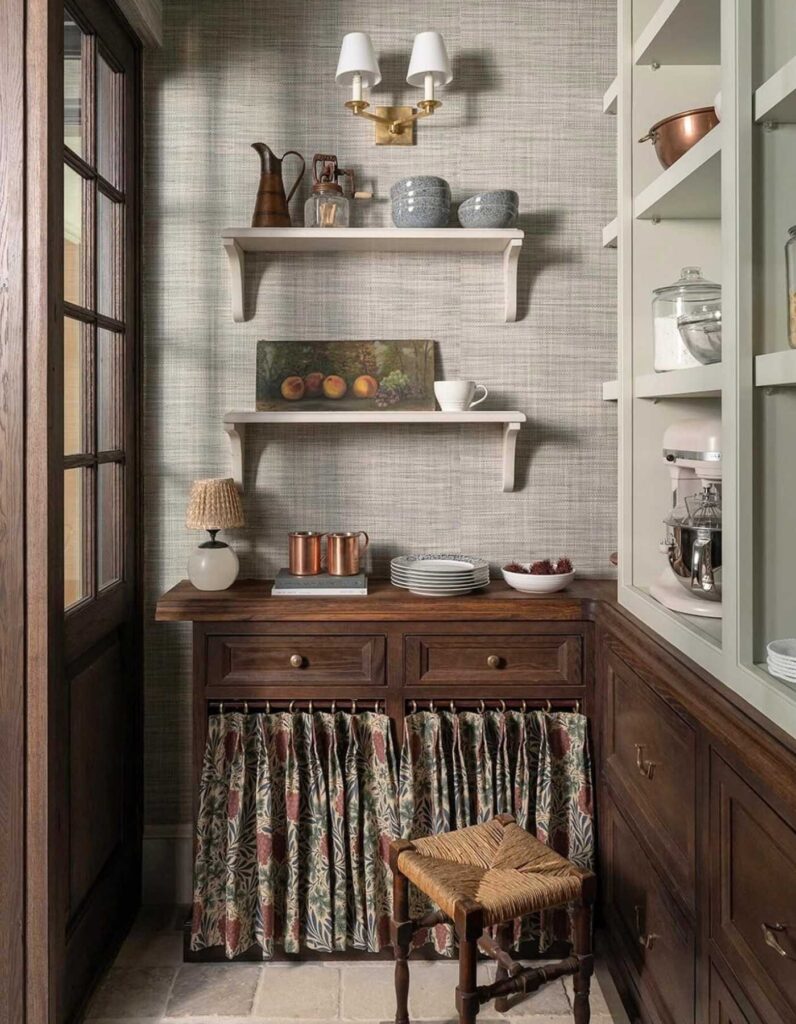 Vintage rustic style farmhouse pantry decorated with shelving, floral cabinet curtains, painting, lamp, and jars.