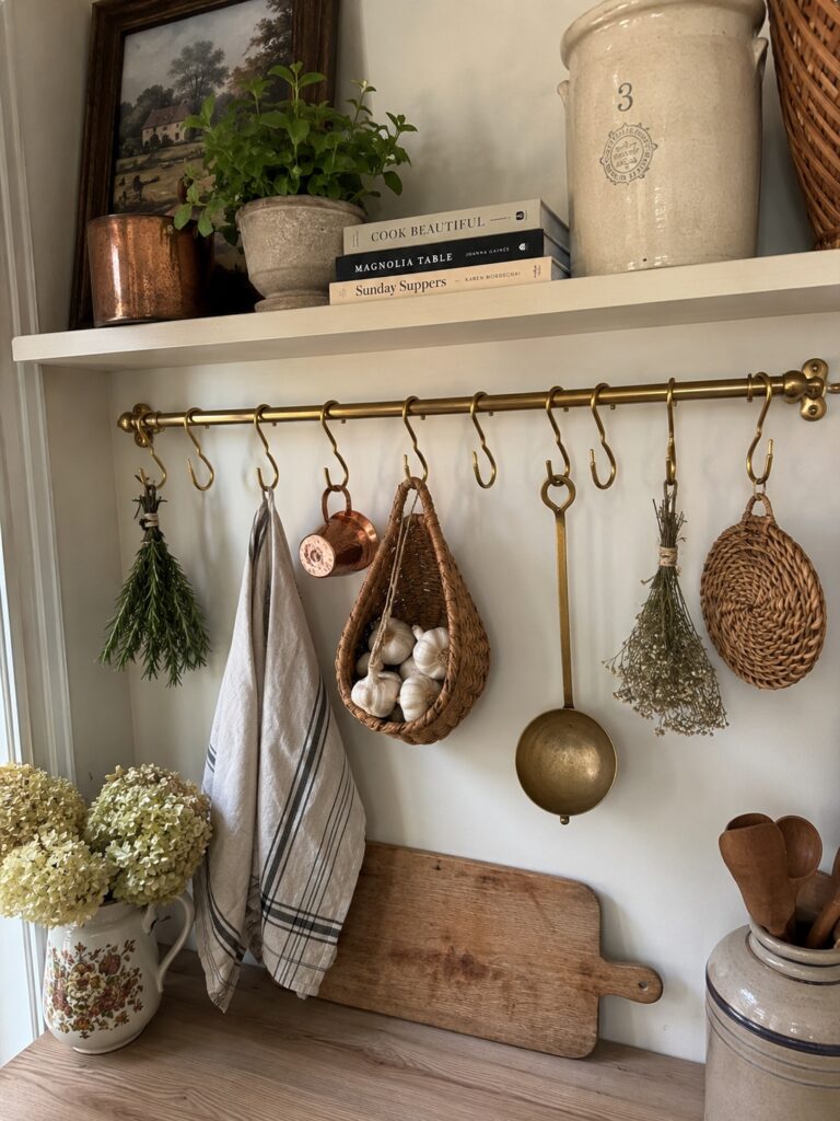 A picture of a country pantry with a gold  hanging rail storage with a tea towel, kitchen utensils, herbs, and other pantry items on the hooks. 