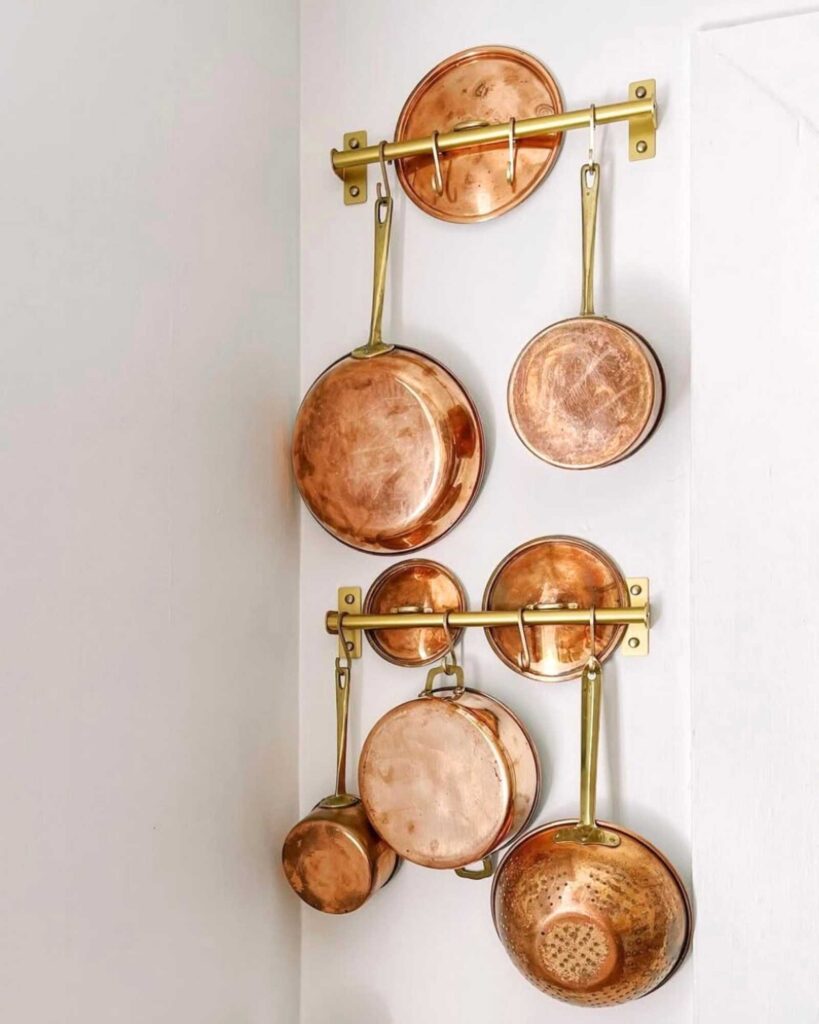 A gold wall-mounted rack with copper pots, pans, and lids. 