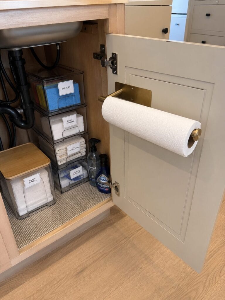 Under kitchen sink organization ideas paper towels on a gold rack behind under sink cabinet door and acrylic bins stacked on top of each other. 