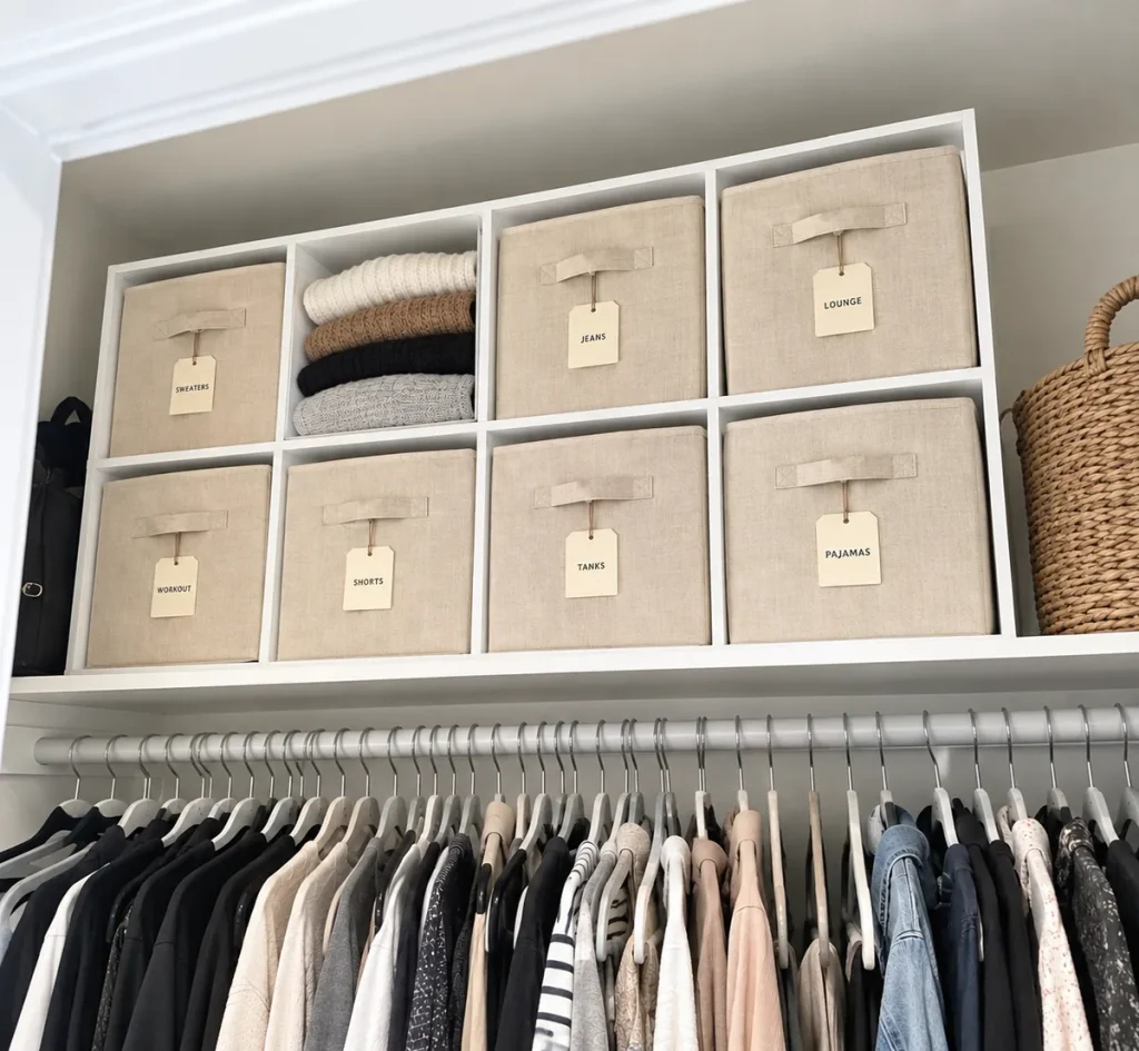Clothes storage ideas: Cube organizer with tan bins on the top closet shelf in a closet with clothes stored inside. 